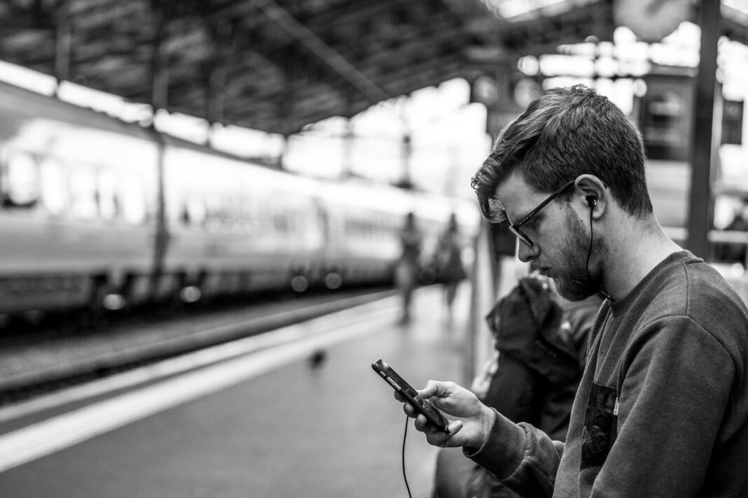 grayscale and selective focus photography of man holding phone at train station